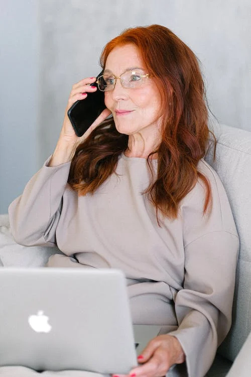 Elderly woman with red hair talking on smartphone at laptop